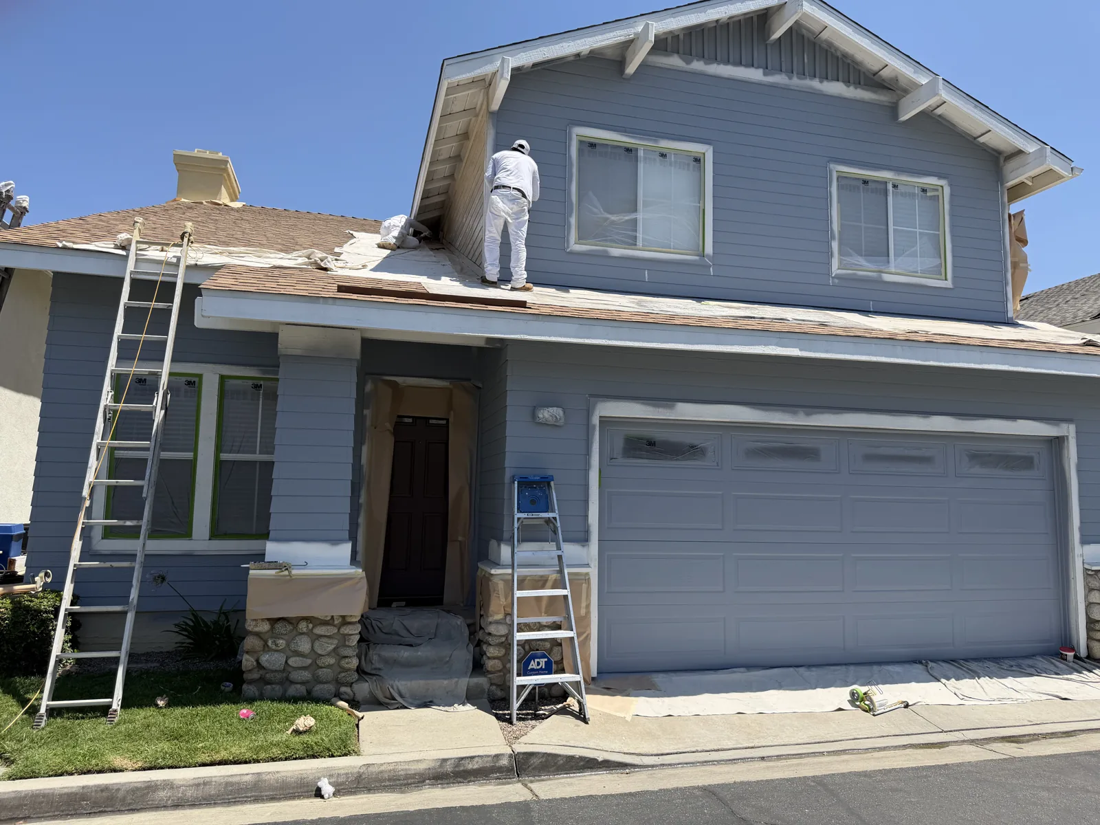 Gray-blue two-story house with white trim, ladders and a painter working near the front porch